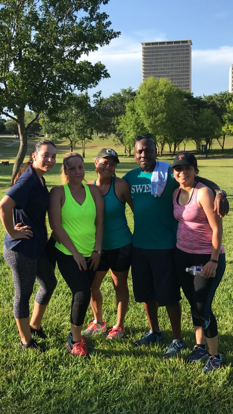 Five people in athletic wear posing together on a grassy park with trees and a tall building in the background