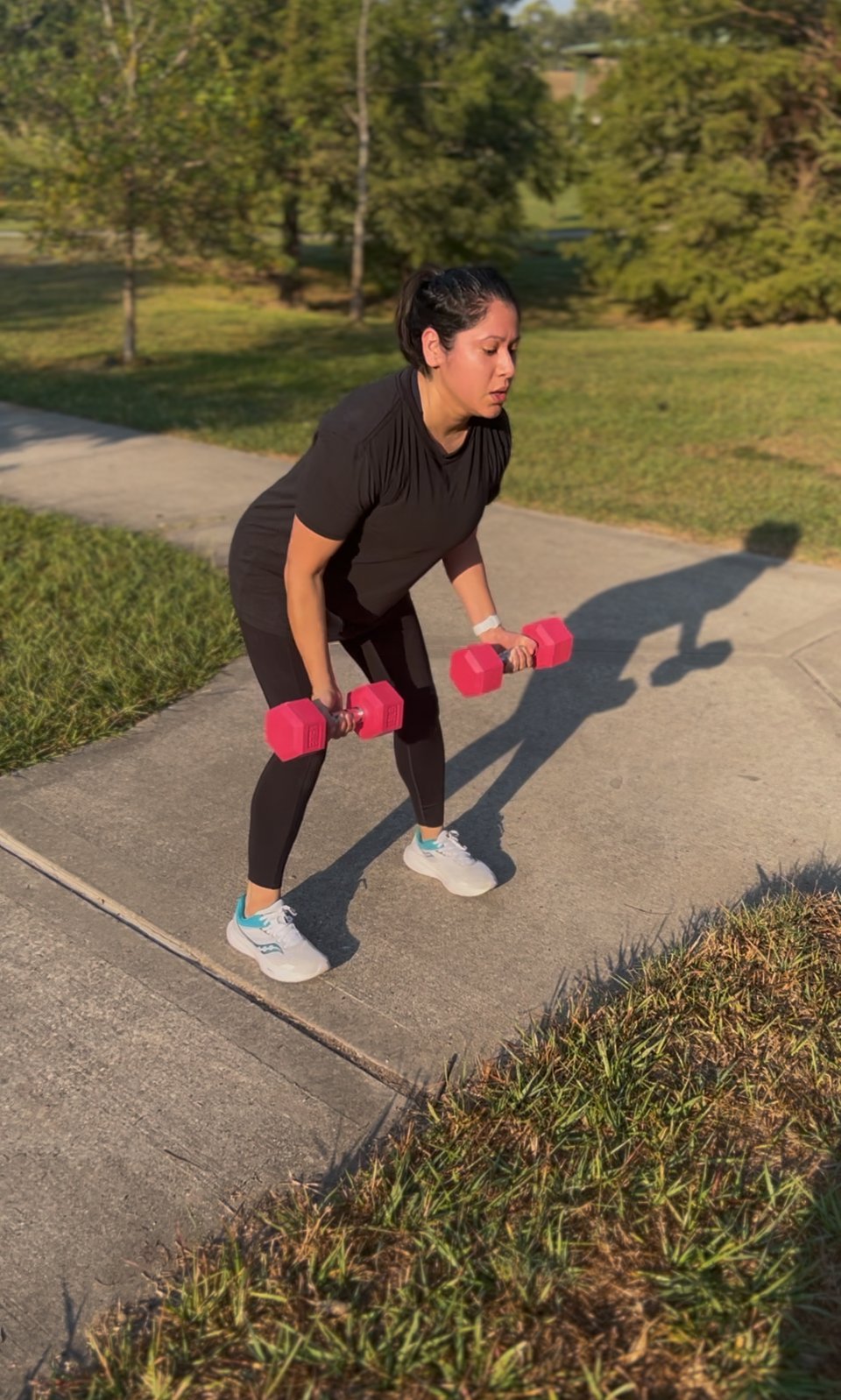 Woman exercising outdoors on a sidewalk holding pink dumbbells in a bent-over position with park scenery in background